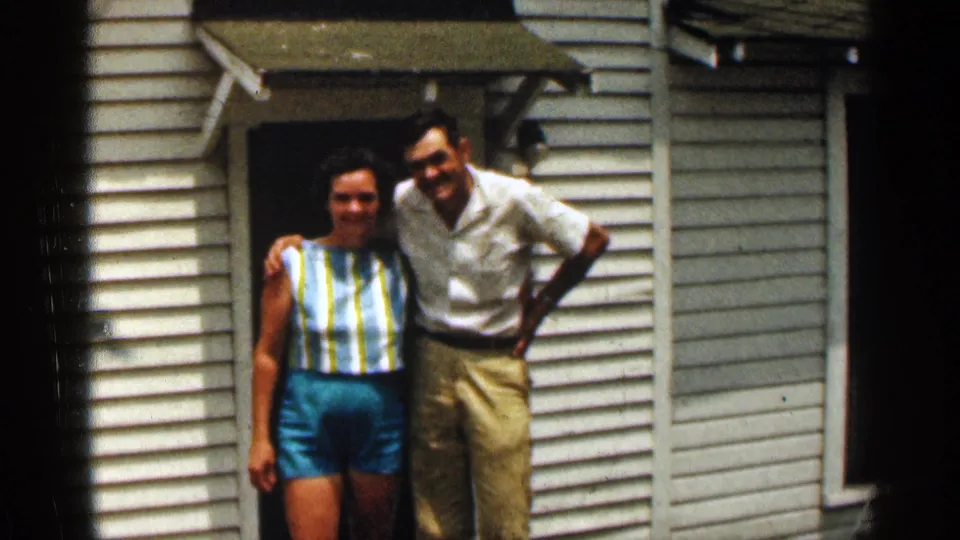 A 1963 Texas home-movie frame shows the couple posing shoulder to shoulder by white siding and a doorway awning.