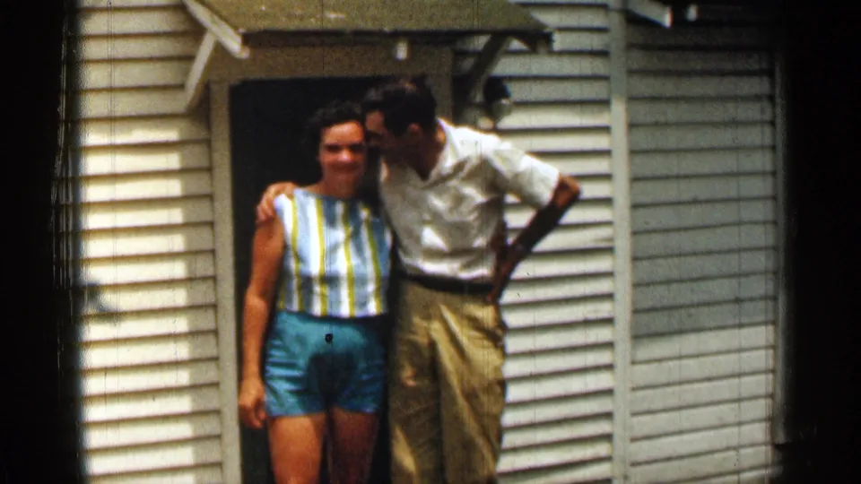 In the final 1963 Texas frame, the man leans toward the woman for a cheek kiss while they stand by the white-sided house.