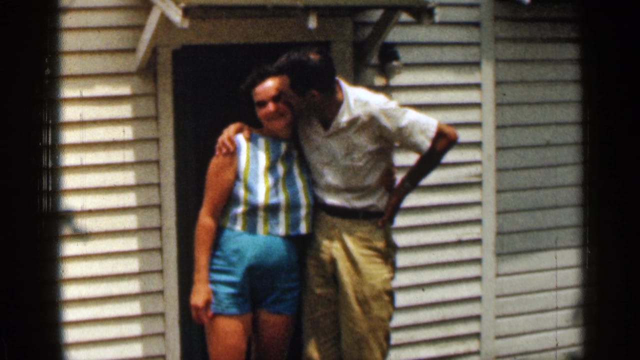 Two adults embrace beside a dark doorway on a white-sided house.