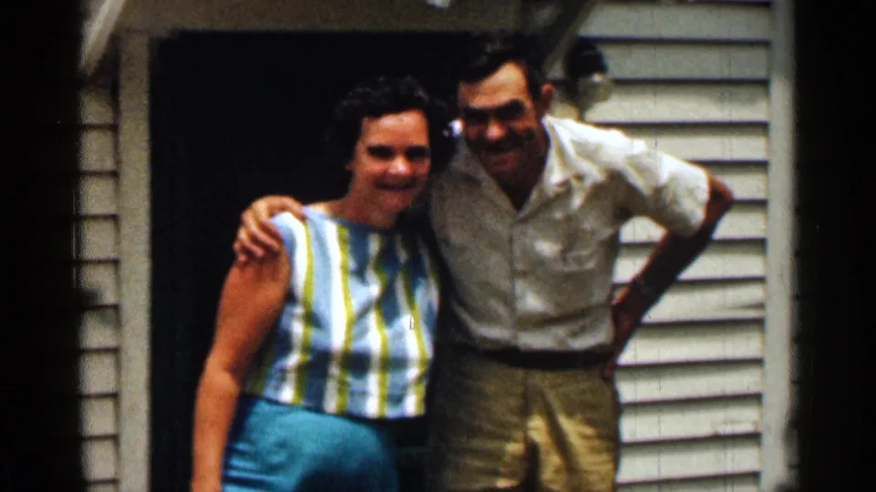 A smiling man and woman pose shoulder to shoulder at a house doorway, with white siding and casual summer clothing visible.