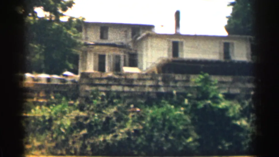 The home-movie view holds on a residential house behind a stone wall, with greenery filling the foreground.