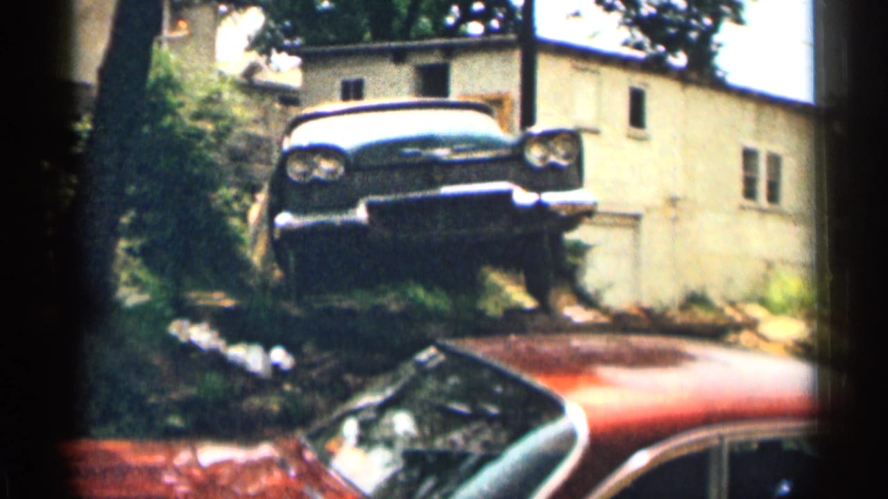 1963 Texas exterior view with a dark car by a residential house and the red body of another parked car crossing the foreground.