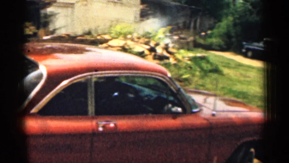1963 Texas home-movie frame showing a red sedan in the foreground, grass, a pale house wall, and a driveway area behind it.