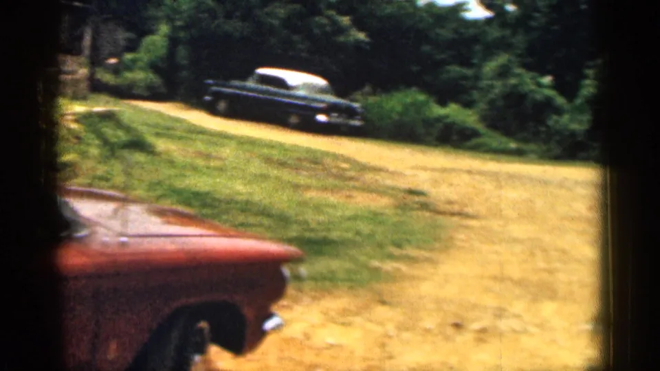 1963 Texas suburban driveway view with a red car in the foreground and a dark sedan parked farther along a dirt path under trees.