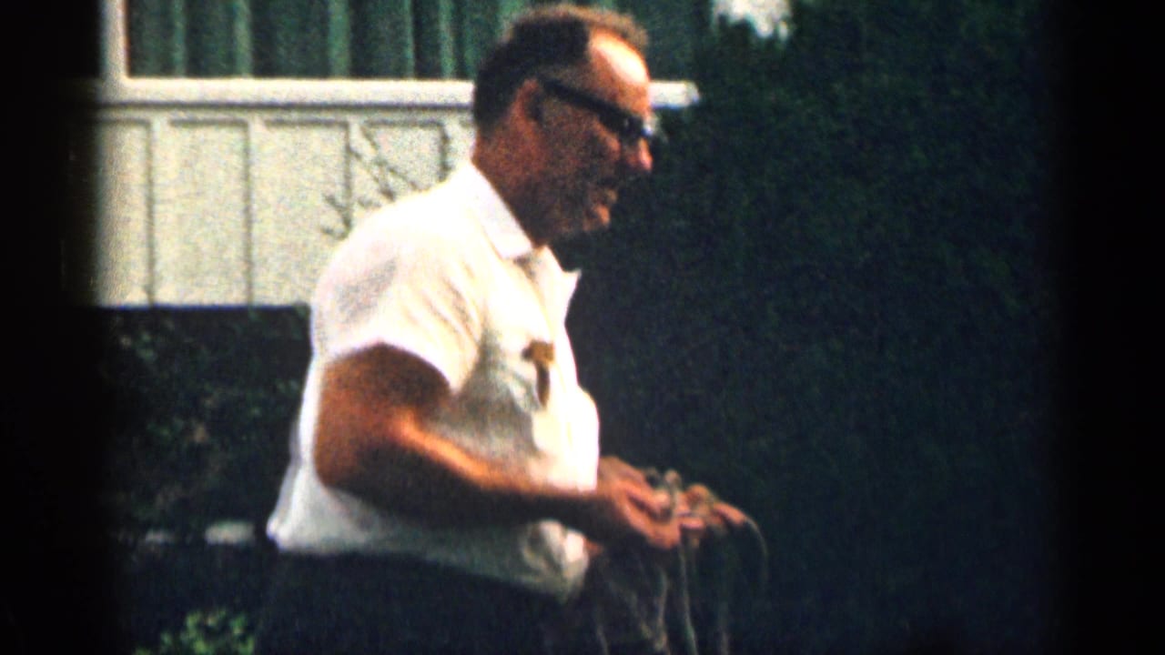 Man in white shirt and dark glasses standing outdoors near a house and greenery.