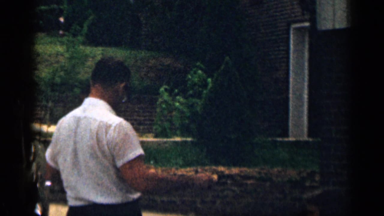 Man seen from behind walking outdoors beside a lawn, shrubs and house exterior.