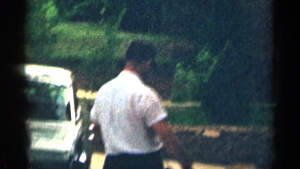 Adult man walks outdoors near shrubs and a partially visible car in 1963 Texas 8mm home-movie footage.