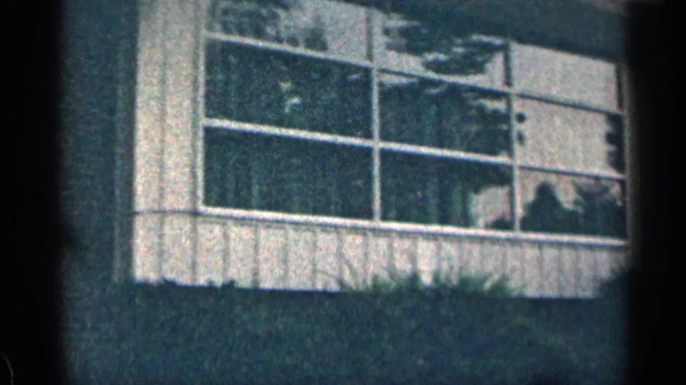 1963 Texas 8mm frame showing a large white-framed window in a brick building, with shrubbery below and reflections across the glass.
