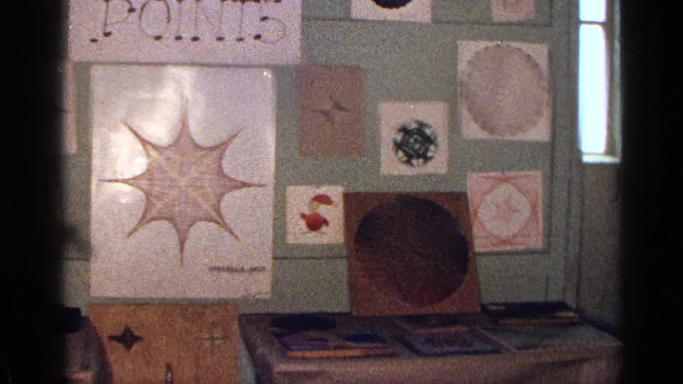1963 Texas home-movie frame showing handmade geometric art and abstract drawings arranged on a green interior wall.