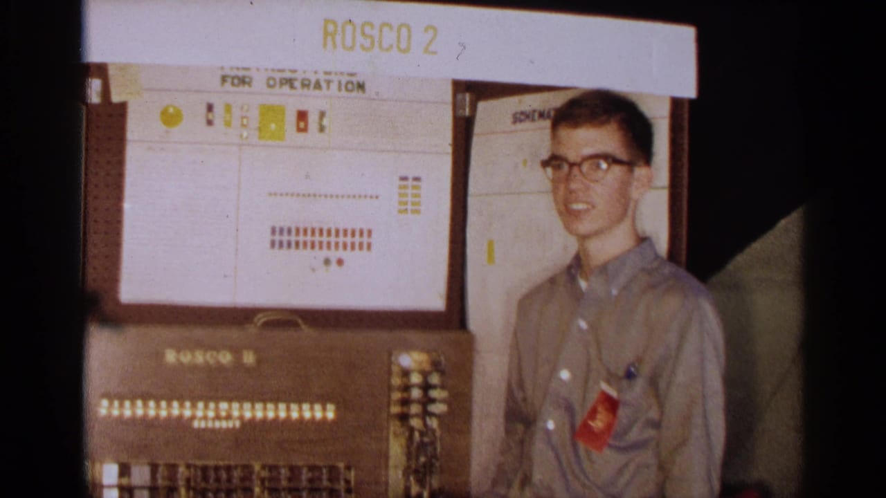 Young man with glasses stands beside a Rosco II console with indicator lights.