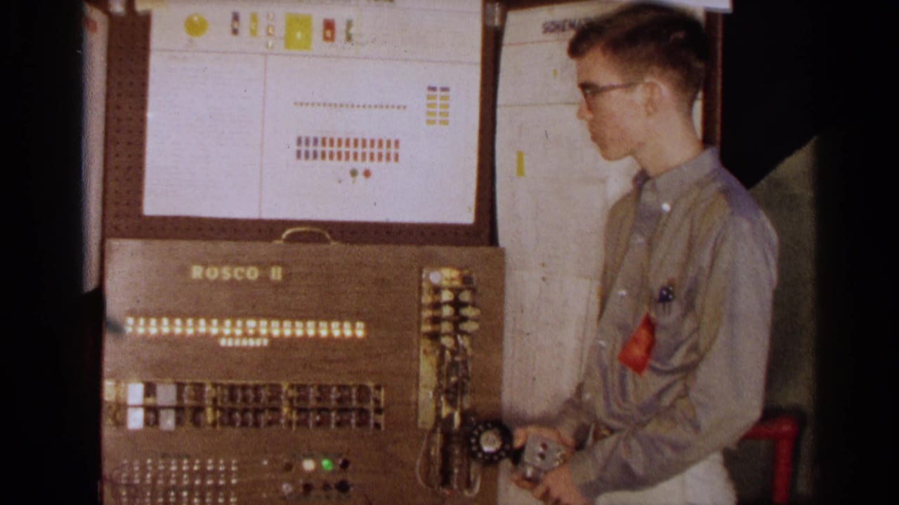 The young man stands in profile while holding controls beside the Rosco II console and wall-mounted operation panel.