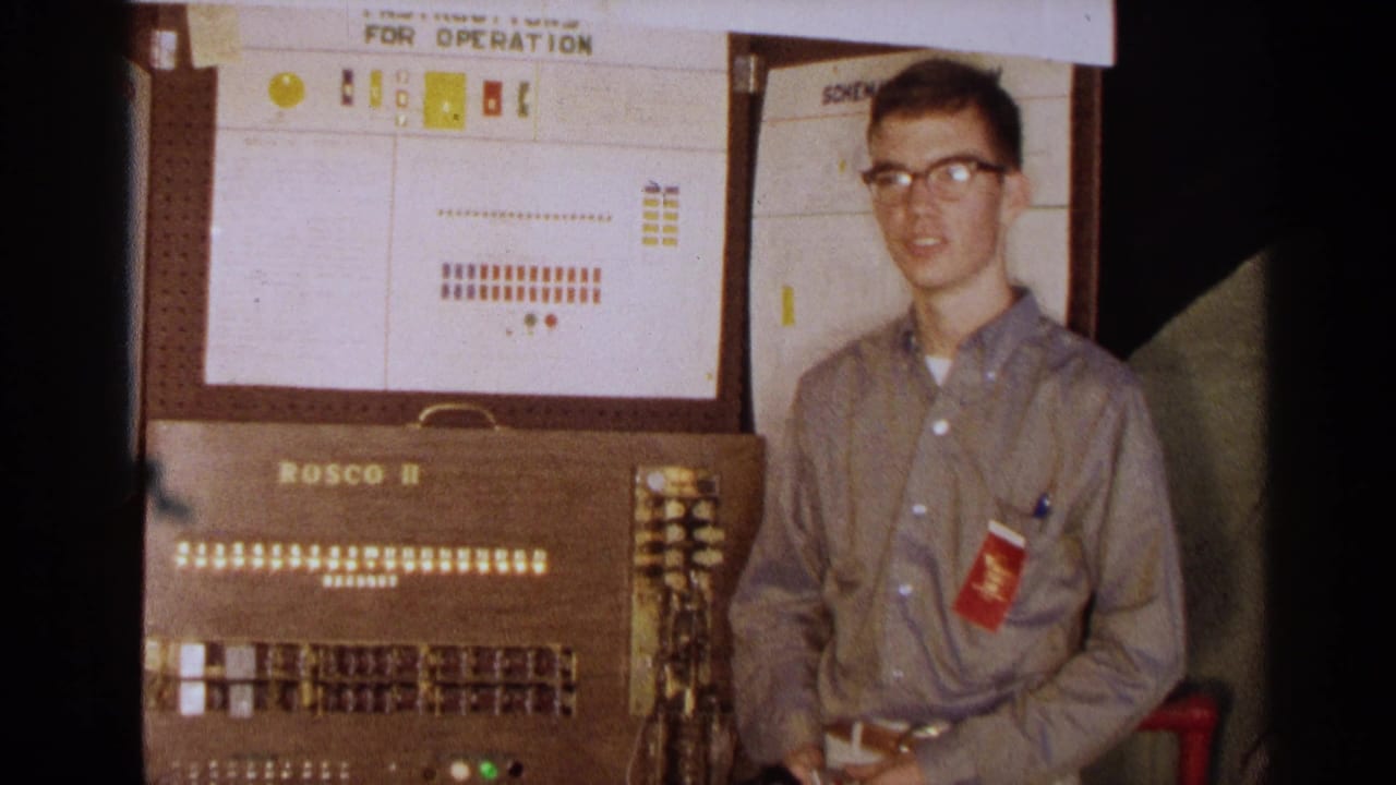 The young man faces the camera beside the Rosco II machine, with rows of switches and small lights visible on the console.