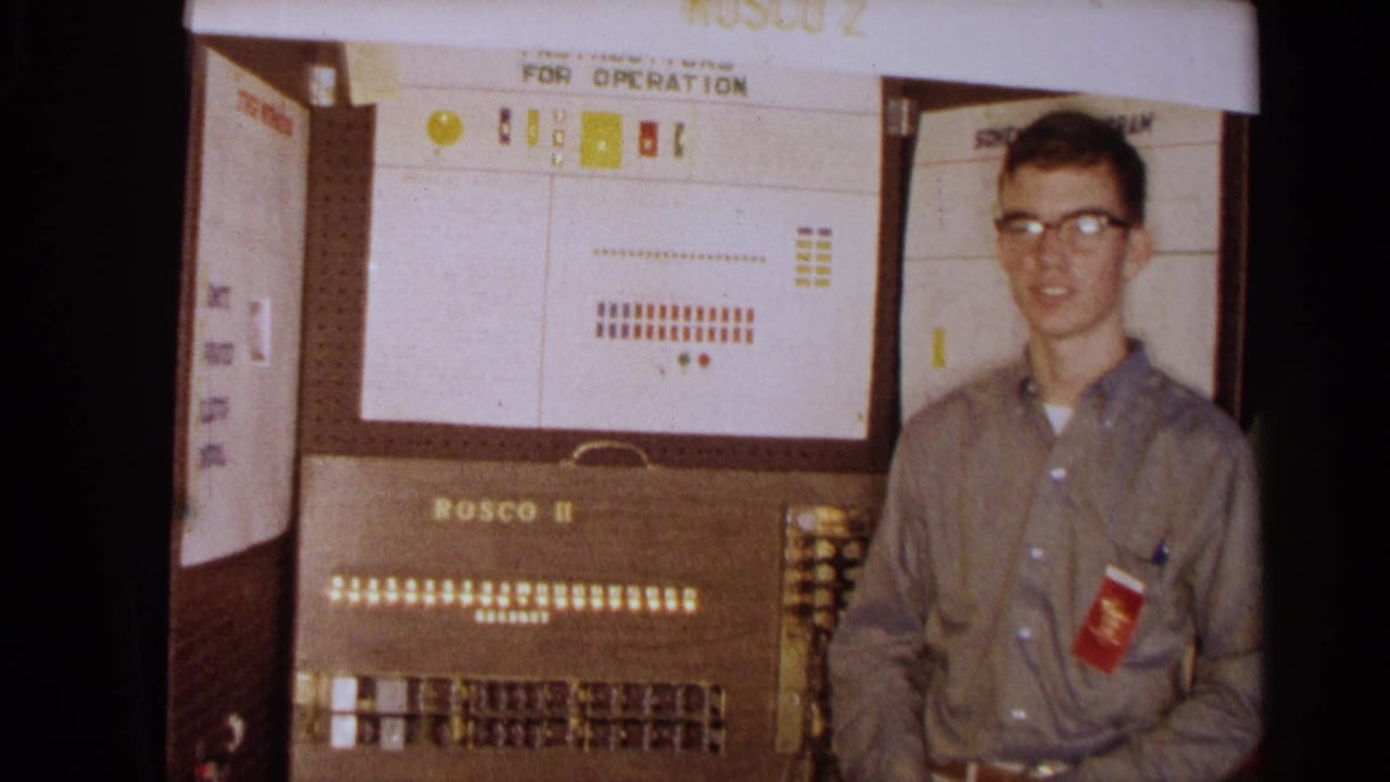 A young man with glasses stands at the side of the Rosco II console, with an operation diagram and illuminated panel behind him.