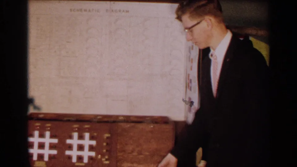 A student in a dark suit stands beside the logic systems project, with illuminated tic-tac-toe grid patterns visible on the base.