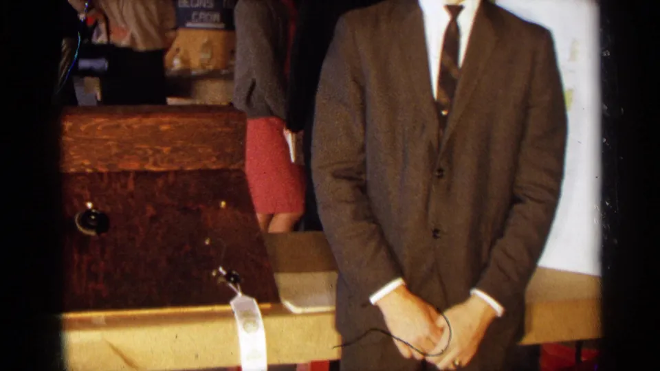 A formally dressed young man stands with hands clasped beside a wooden apparatus at a 1960s Texas school science display.