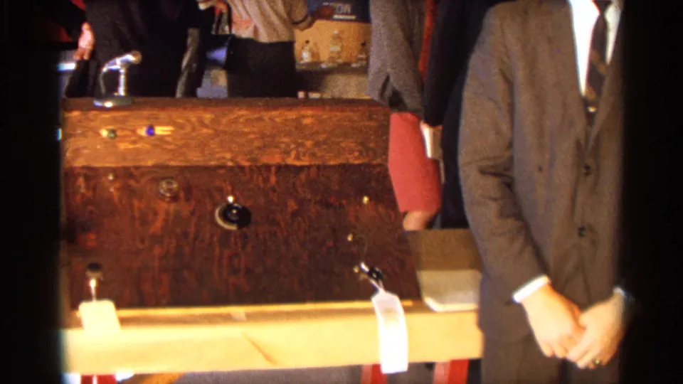 The final frame shows the wooden apparatus in front of the suited presenter, with neighboring fair displays partly visible behind.