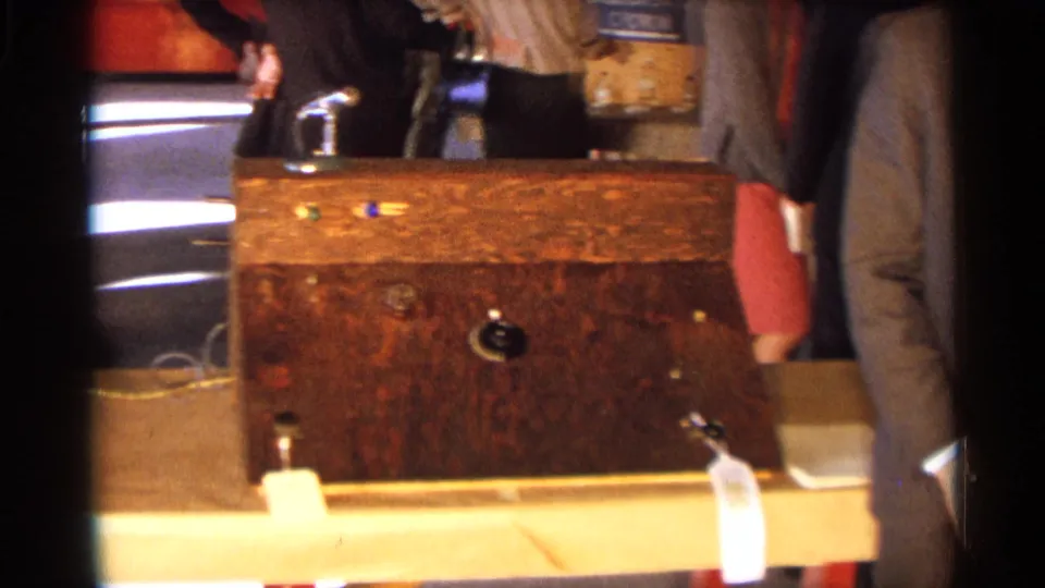 1963 Texas home-movie frame showing a wooden science display case with a small rocket model and adults standing nearby.