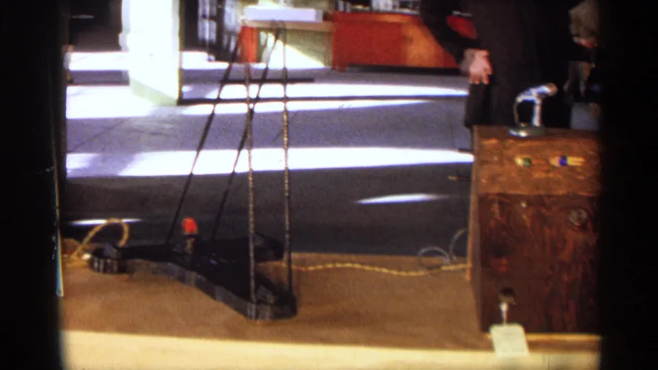 1963 Texas home-movie frame with a tripod stand, floor cables, and part of a wooden science fair display case.