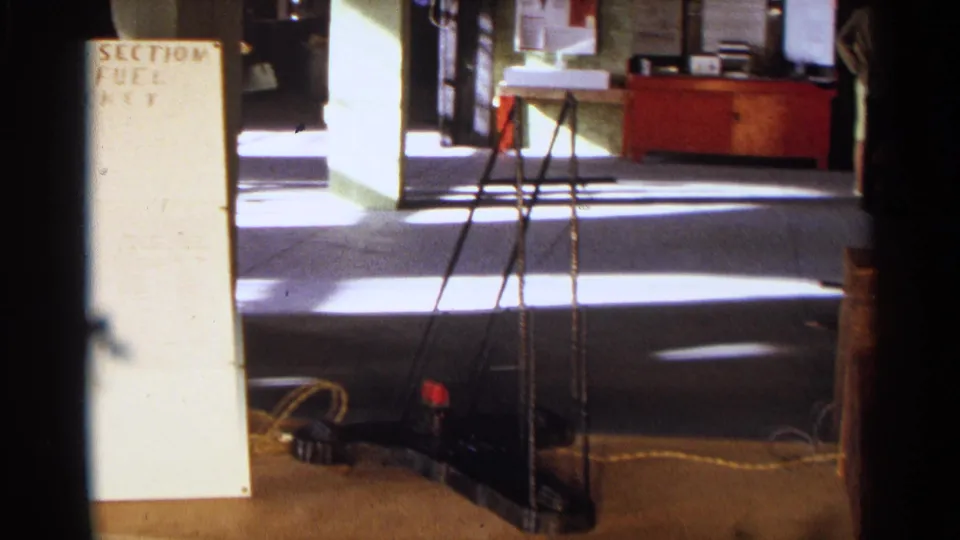 1963 Texas home-movie frame showing a science display board at left, with tripod legs, cables, and room furnishings behind it.