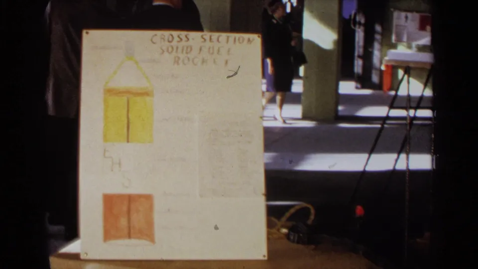 1963 Texas home-movie frame showing a hand-drawn solid fuel rocket cross-section board and visitors moving through the room.