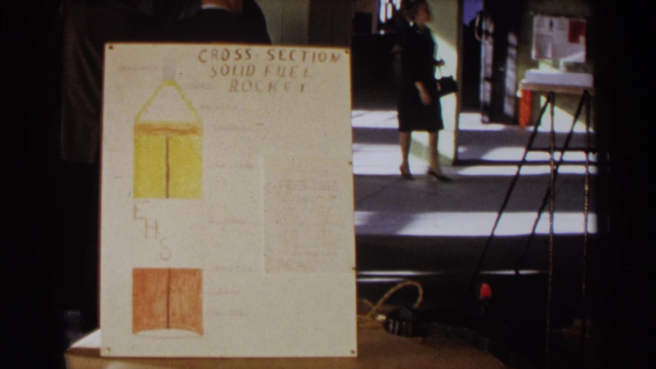 1963 Texas 8mm frame showing a hand-lettered solid fuel rocket cross-section board with hallway visitors behind it.