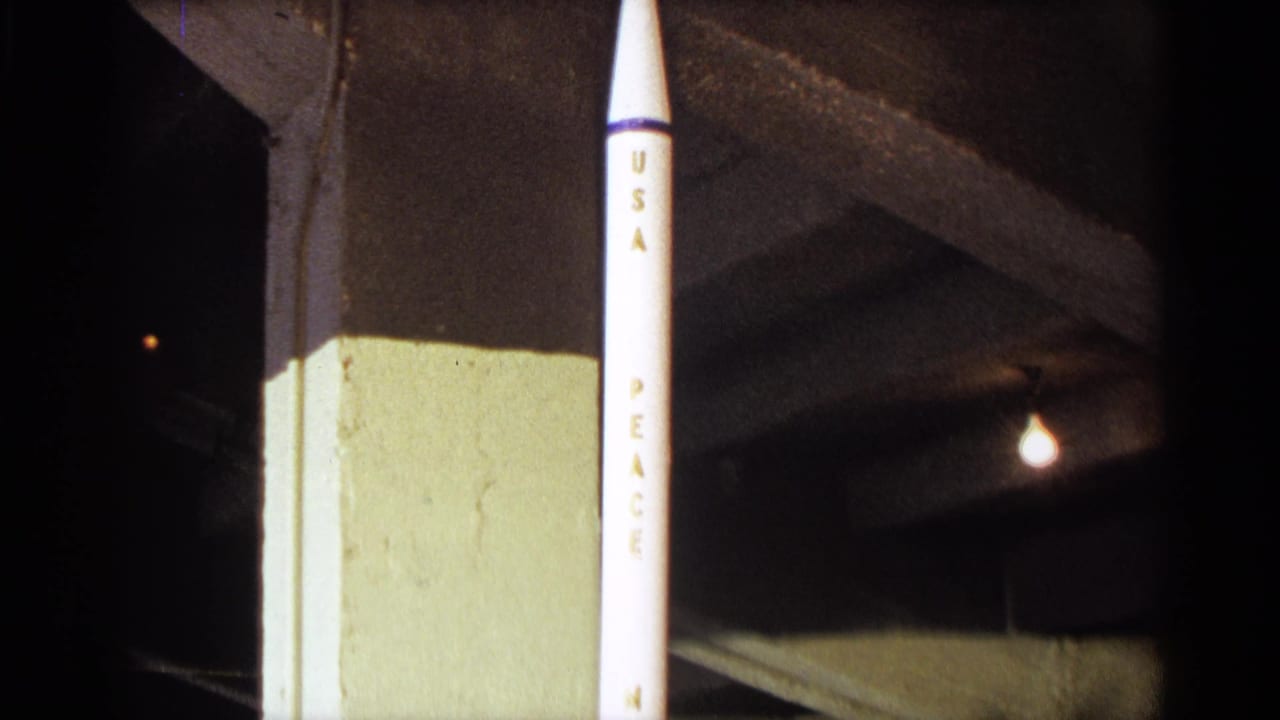 1963 Texas home-movie frame showing a white model rocket marked with vertical lettering near a concrete column and overhead light.