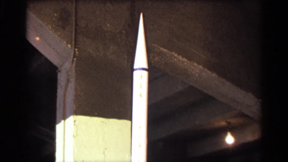 Close 8mm view of a white model rocket in an indoor exhibition area, with a concrete support and hanging light visible behind it.