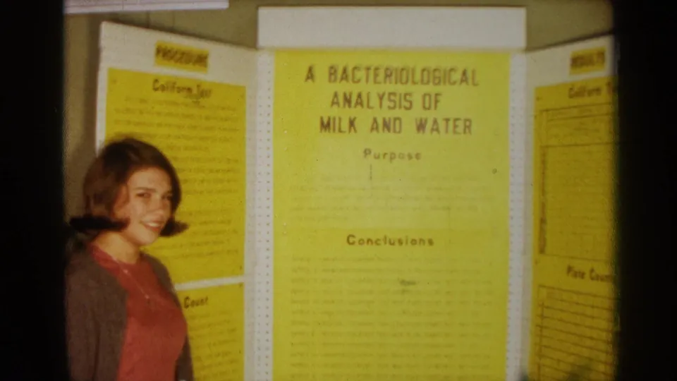 Young woman at a 1963 Texas science fair display titled A Bacteriological Analysis of Milk and Water, filmed in warm 8mm color.