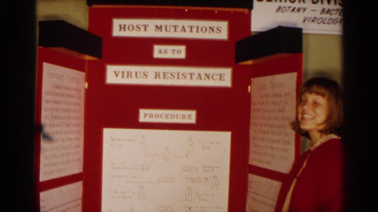 In 1963 Texas home-movie footage, a young woman stands beside a red tri-fold display titled Host Mutations As To Virus Resistance.
