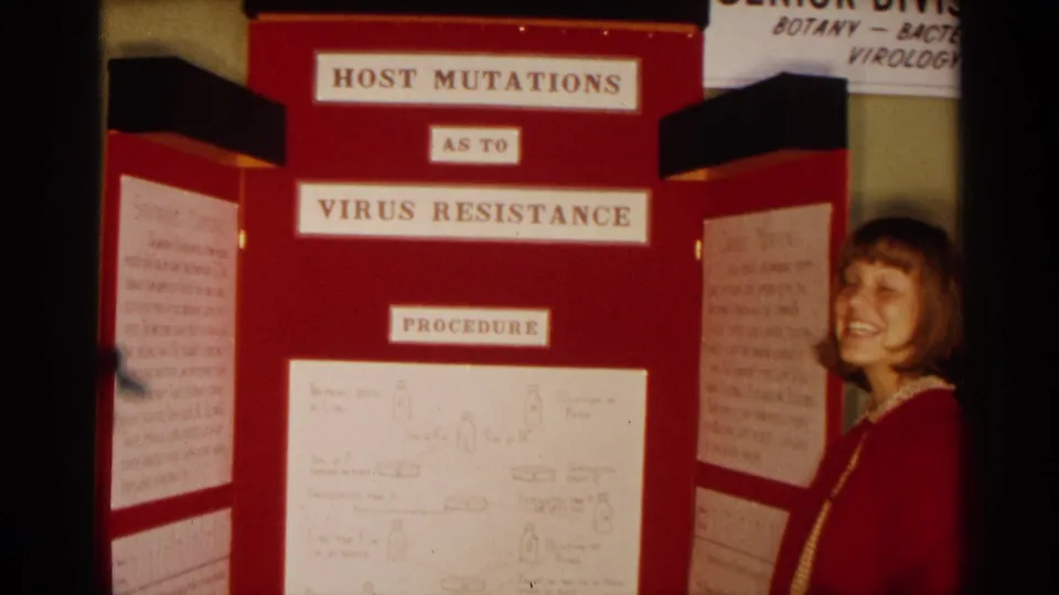 In 1963 Texas home-movie footage, a young woman stands beside a red tri-fold display titled Host Mutations As To Virus Resistance.