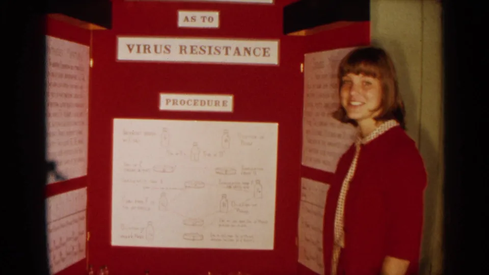 The presenter smiles beside the red virus resistance display, with typed labels and handwritten science notes visible on the board.