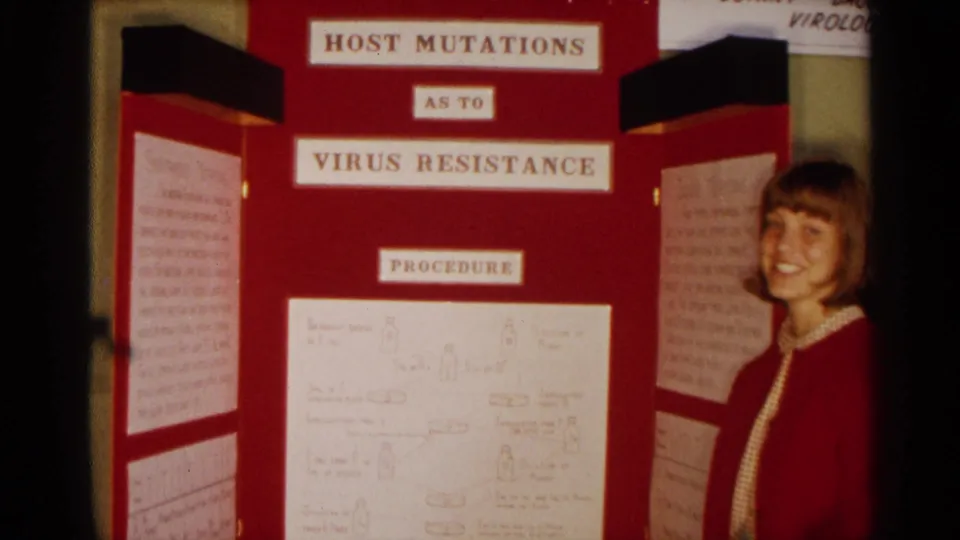 A young woman in a red cardigan poses beside the Host Mutations As To Virus Resistance board in the 1963 Texas science fair footage.