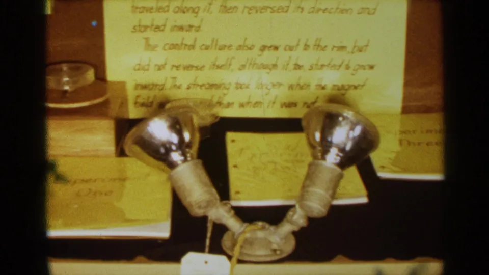 1963 Texas home-movie frame showing the lower section of the cytoplasmic streaming display, with handwritten notes, a petri dish, paired metal lamp heads, a diagram page, and labels arranged on the table.