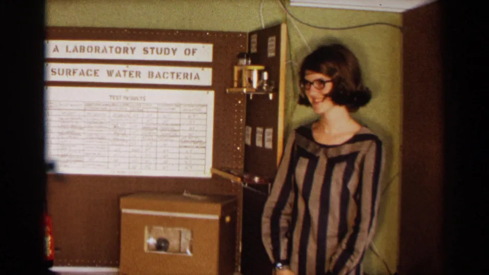 Young woman in a striped dress and glasses stands by a 1963 Texas science fair display titled as a laboratory study of surface water bacteria.