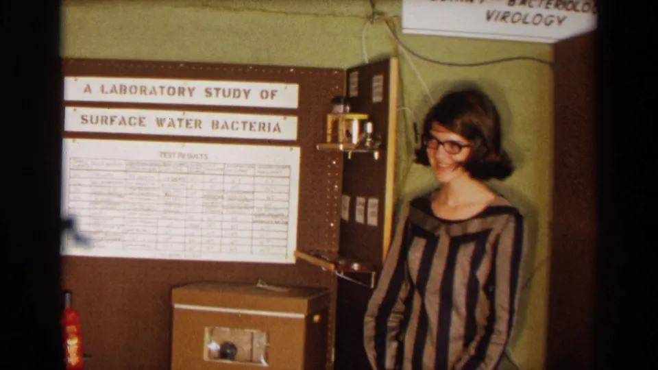 In 1963 Texas home-movie footage, a young woman in glasses and a striped dress stands beside a school display titled "A Laboratory Study of Surface Water Bacteria."