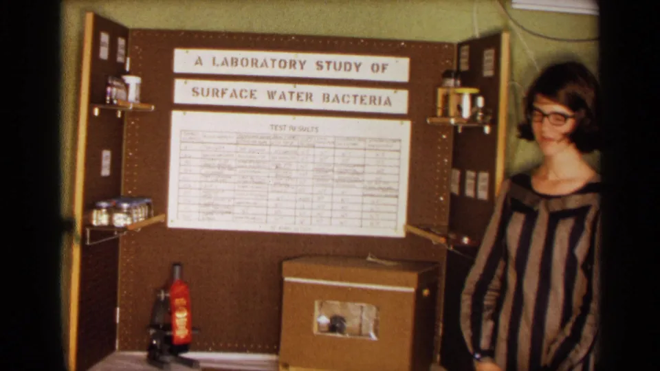 The student presenter smiles near a handmade display board for a surface water bacteria study, with a microscope, sample bottles, and a large test-results sheet visible.