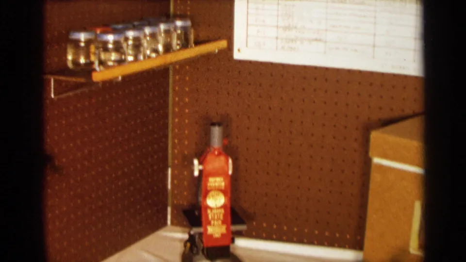1963 Texas 8mm frame showing a lab display board with glass jars, pinned notes, and a red wall-mounted object below the shelves.