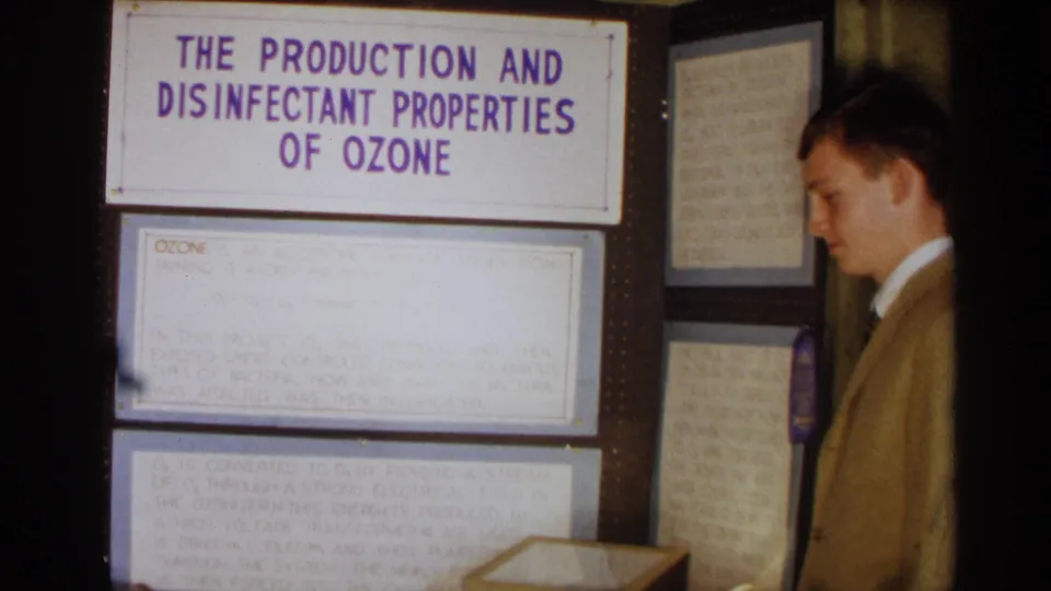 1963 Texas 8mm frame showing the ozone science project board, notes, and a suited young man in profile.