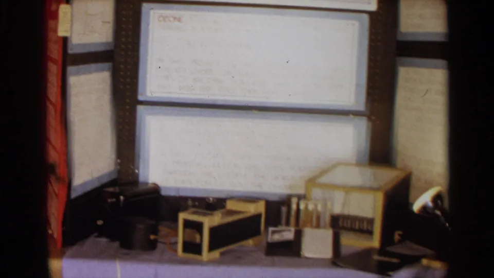 1963 Texas 8mm frame showing a high school science fair ozone display with handwritten boards and small equipment on a purple-covered table.