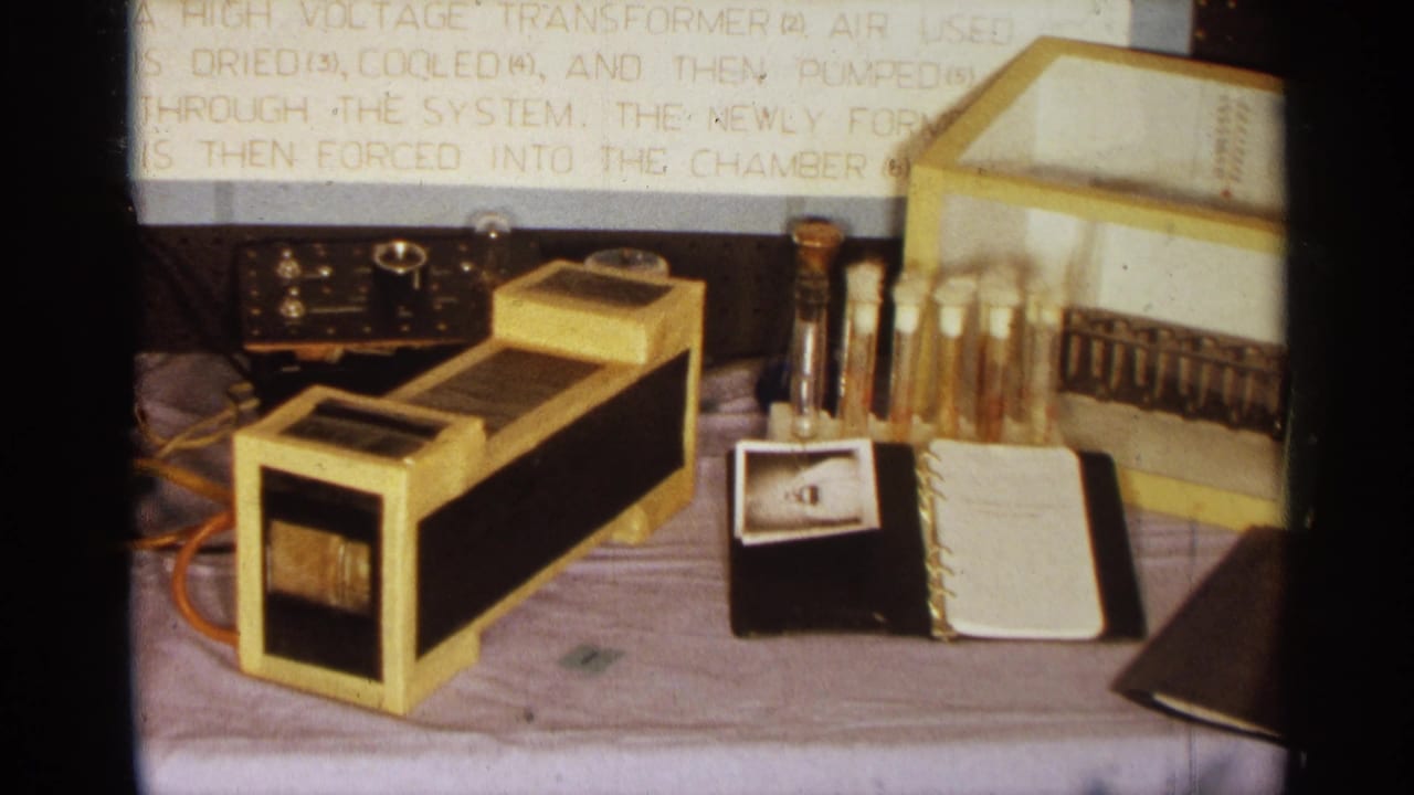 1963 Texas 8mm frame showing a tabletop science demonstration with a gold transformer, analog meter, test tubes, notebook, and display text.