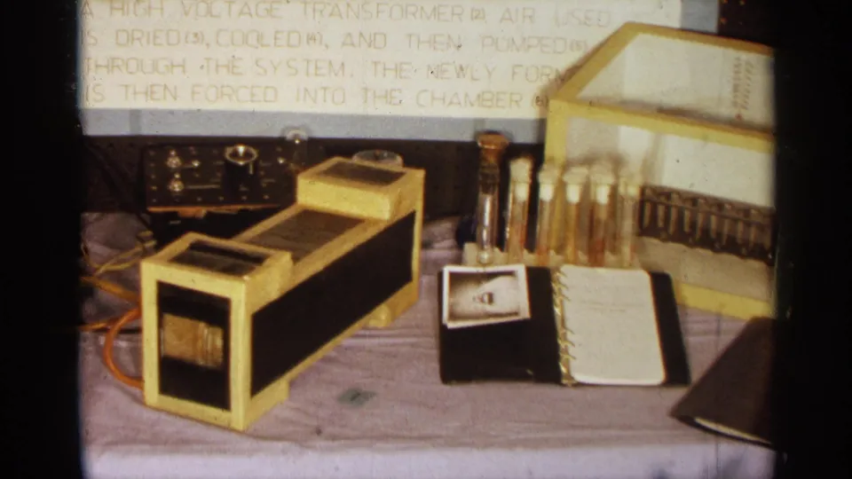 1963 Texas 8mm frame showing a tabletop science demonstration with a gold transformer, analog meter, test tubes, notebook, and display text.