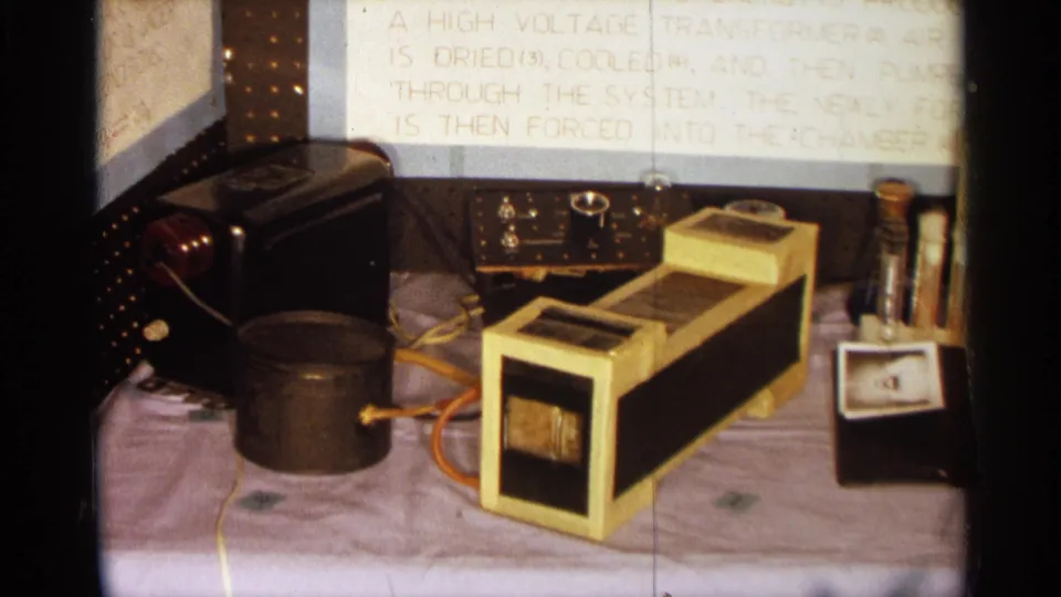 1963 Texas home-movie frame of a science demonstration table with transformer, analog meter, wires, and a partially readable display board.