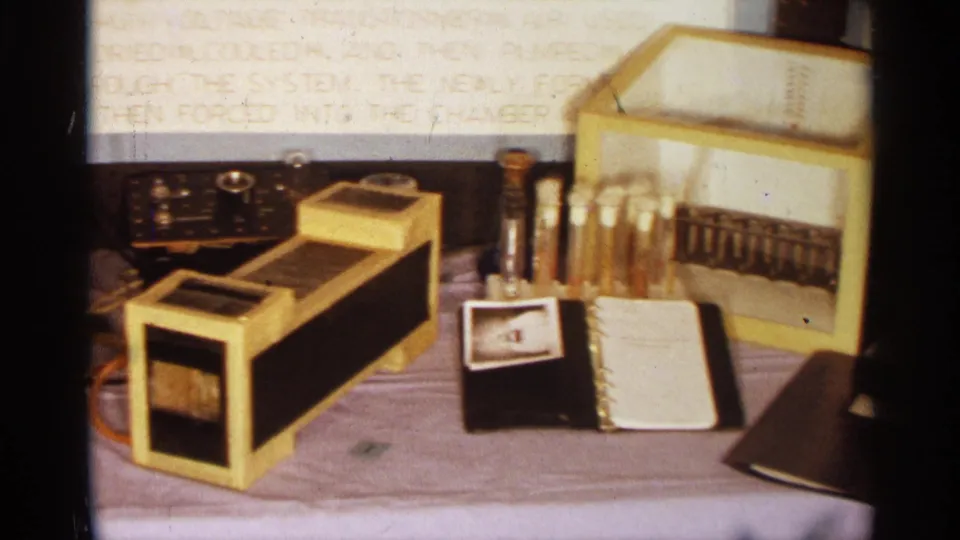 1963 Texas 8mm frame showing the transformer and adjacent analog meter, test tubes, notebook, wires, and handwritten demonstration text.