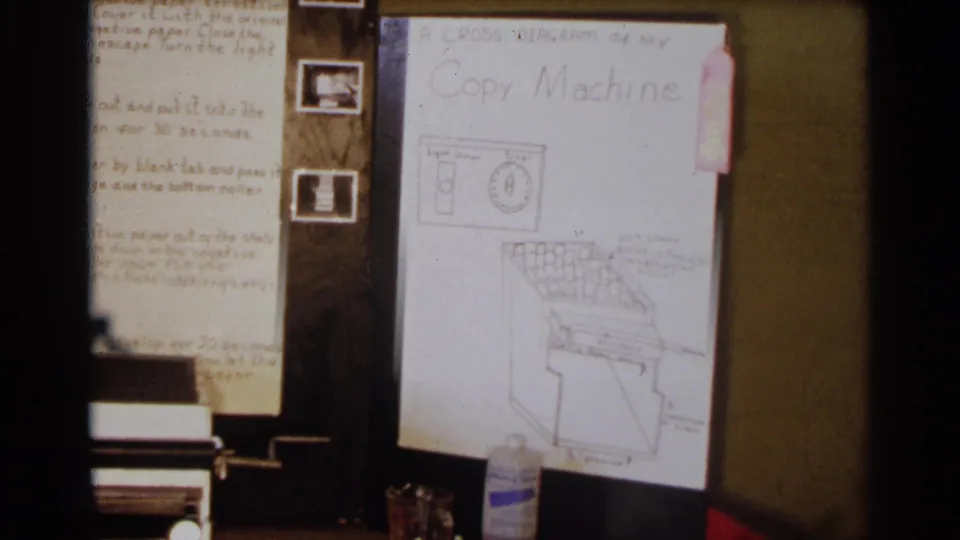 1963 Texas 8mm frame showing a wooden science display with handwritten notes, a copy-machine diagram, and small equipment on a table.