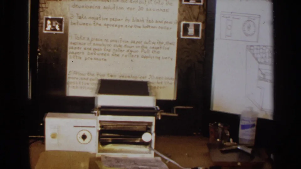 White tabletop apparatus sits before handwritten photographic-development notes on a hinged wooden display in 1963 Texas footage.