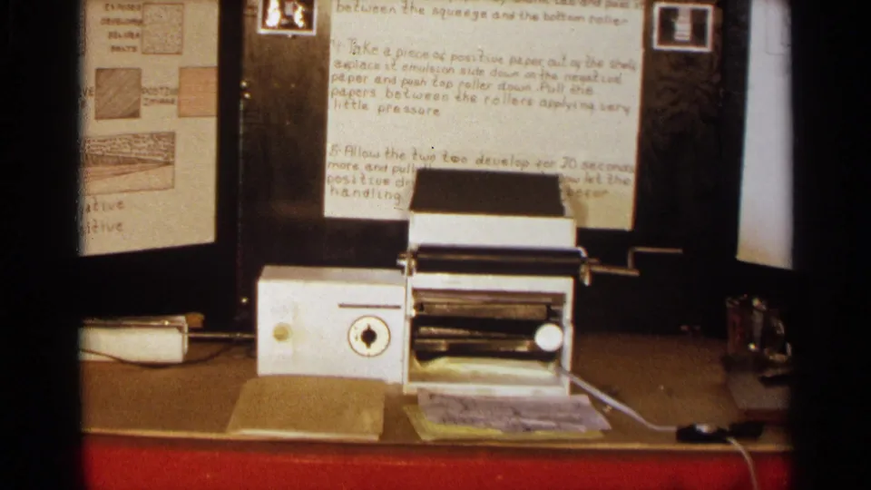 8mm frame of a student science display with notes, a small white machine, loose paper, and a bottle near the right edge.