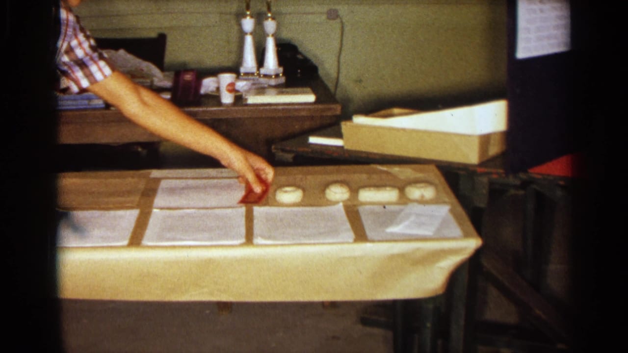 1963 Texas 8mm frame showing a partial person reaching over a beige-covered table with typed papers and small white objects.