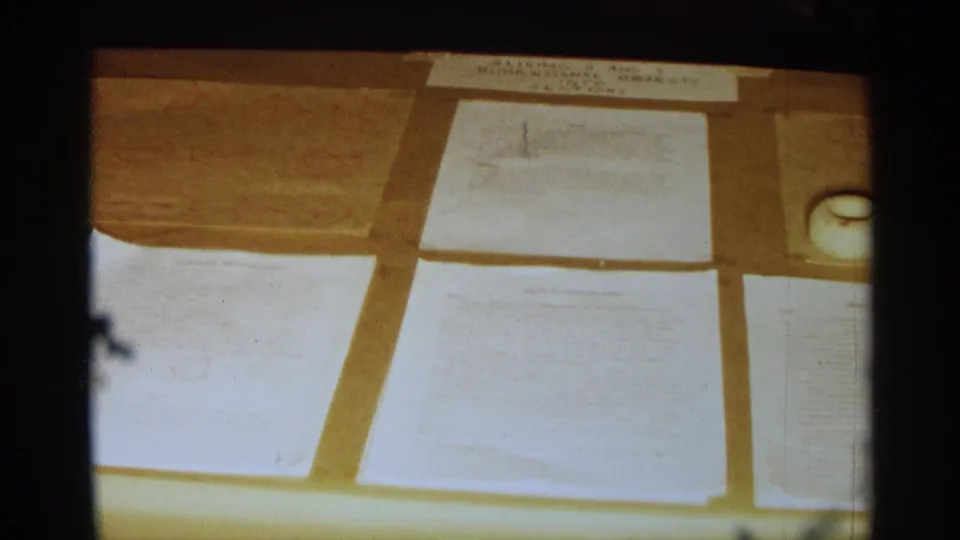 1963 Texas home-movie close-up of papers spread on a beige table covering with white round objects at the side.