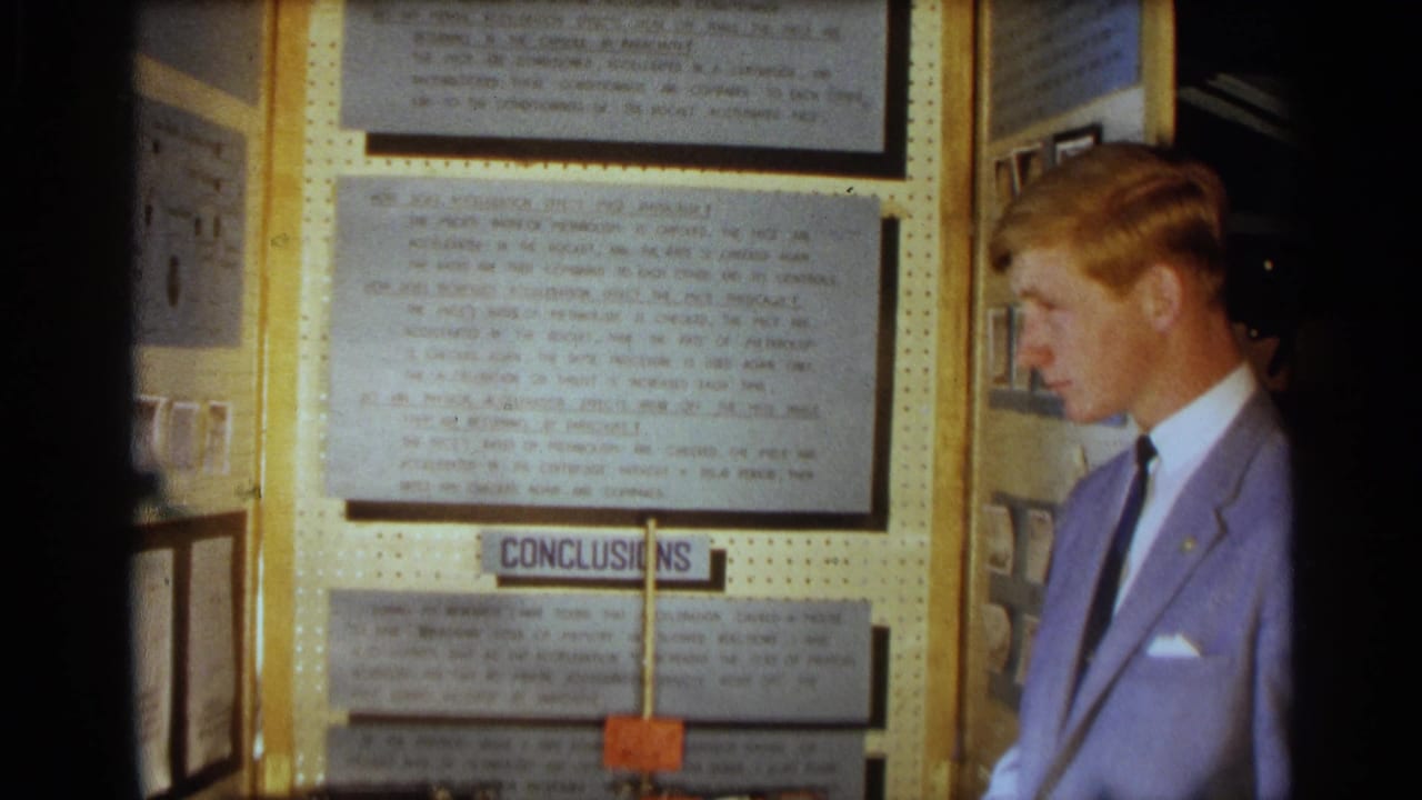 Young man in a light blue suit stands beside a science fair display board with typed panels.