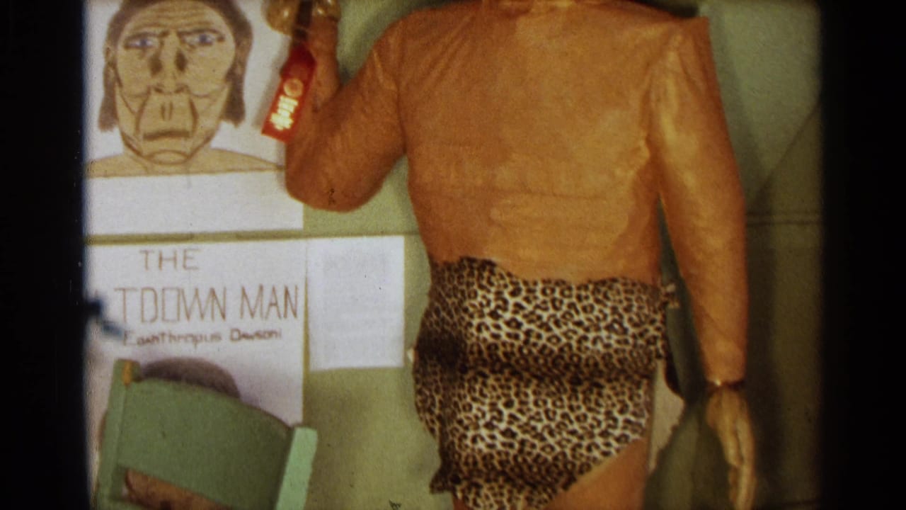 Caveman statue torso in leopard-print loincloth beside a green chair and evolution drawings.